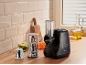 Black electric slicer with cutting and grating attachments, surrounded by fresh vegetables.