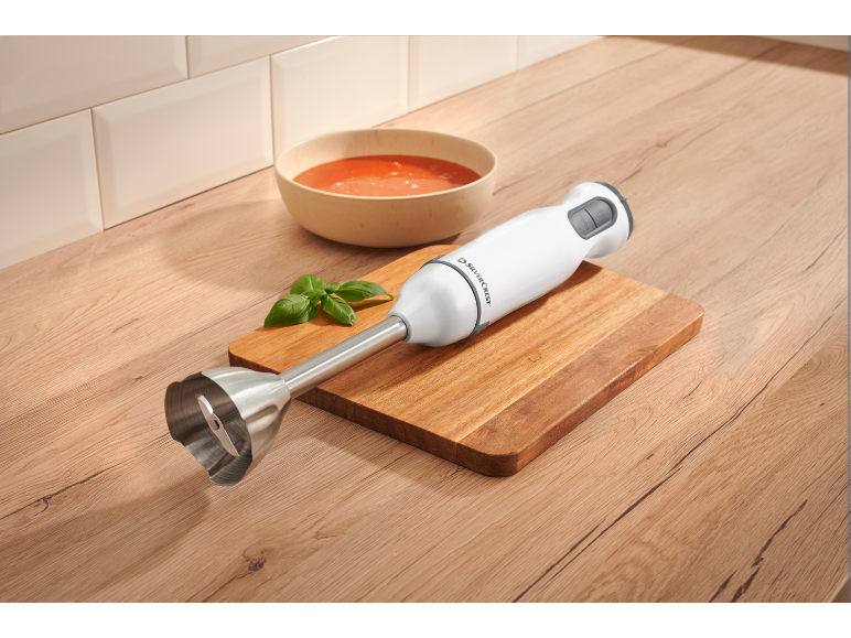 Immersion blender on a wooden board next to a bowl of tomato soup and basil.