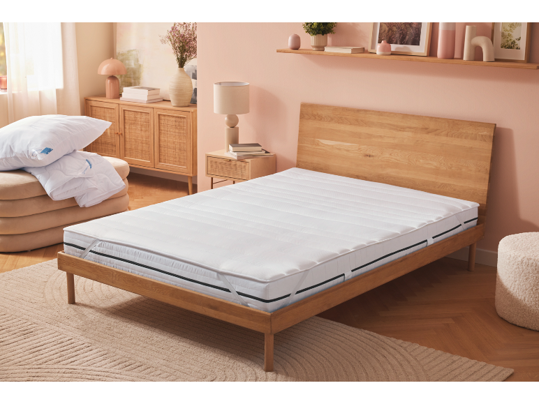 Bedroom with white mattress, pillows and duvet on a pouf, and wooden furniture.