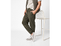 Man in olive green joggers, beige t-shirt, and quilted vest, leaning on a chair.