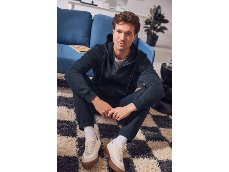 Man in dark blue tracksuit and white sneakers sitting on a rug.