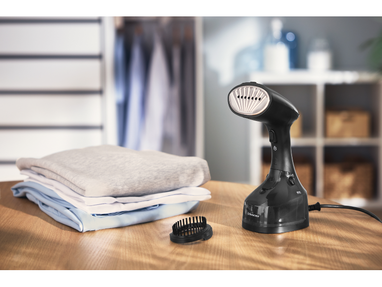 Black garment steamer with accessories and folded clothes on a wooden table.
