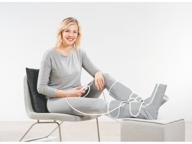 Woman relaxes in chair wearing leg massage boots, holding a control panel.