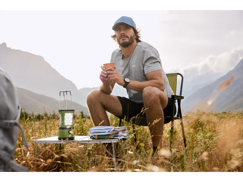 Man in cap and t-shirt sits on folding chair with mug, next to a camping lantern.
