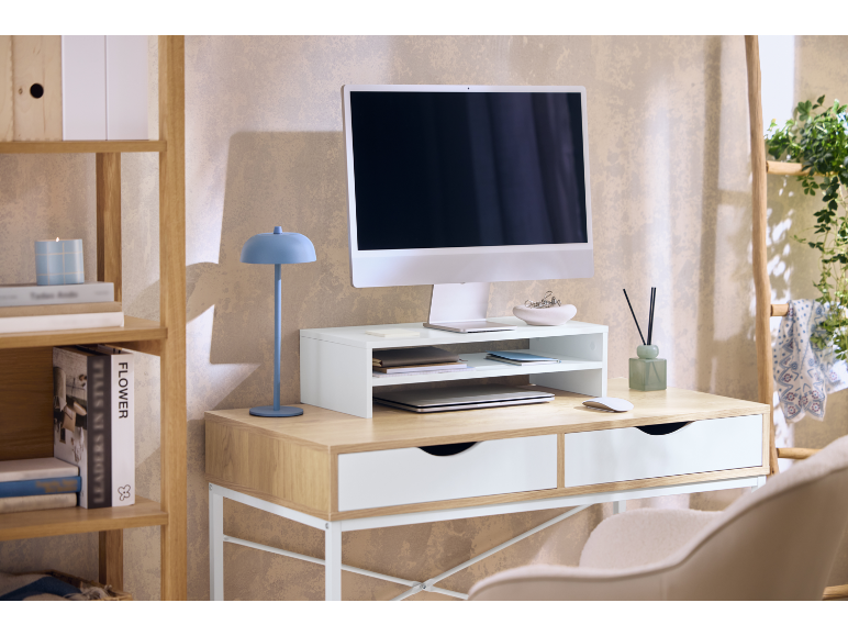 Home office desk with a computer, monitor stand, blue lamp, and wooden shelving.