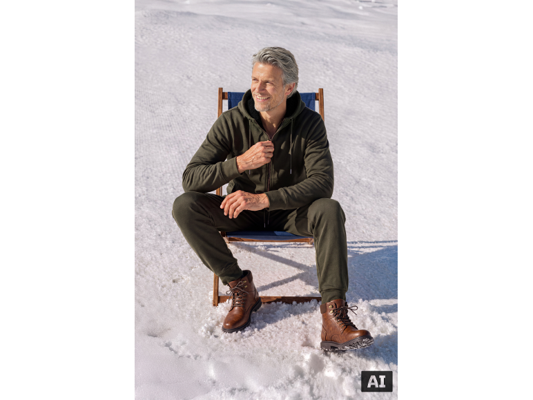 Man in green tracksuit and brown boots sitting on a chair in the snow.