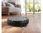 Black robotic vacuum cleaner on a wooden floor with dirt, with a sofa and window in the background.