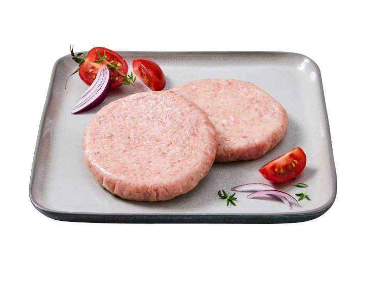 Two raw chicken burgers on a plate, garnished with tomato, onion, and herbs.