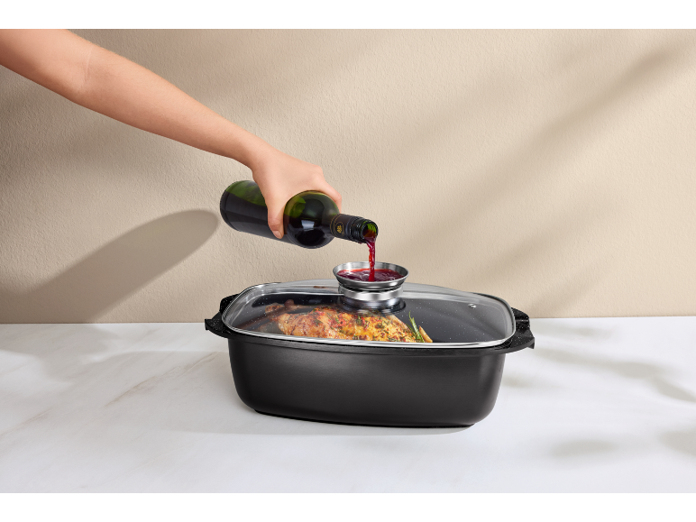 A hand pours red wine from a bottle into a roasting pan with a roasted chicken.