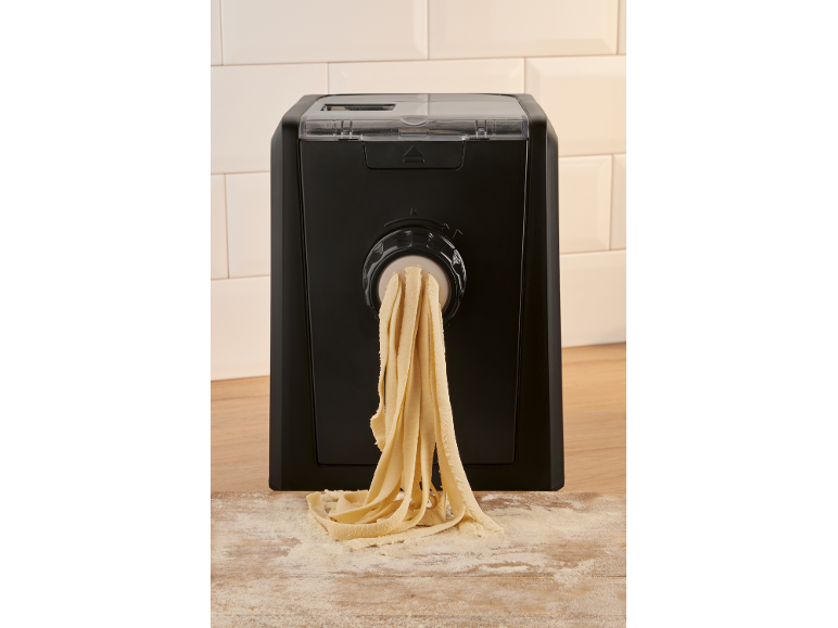 Fresh pasta being extruded from a black pasta maker onto a floured wooden surface.