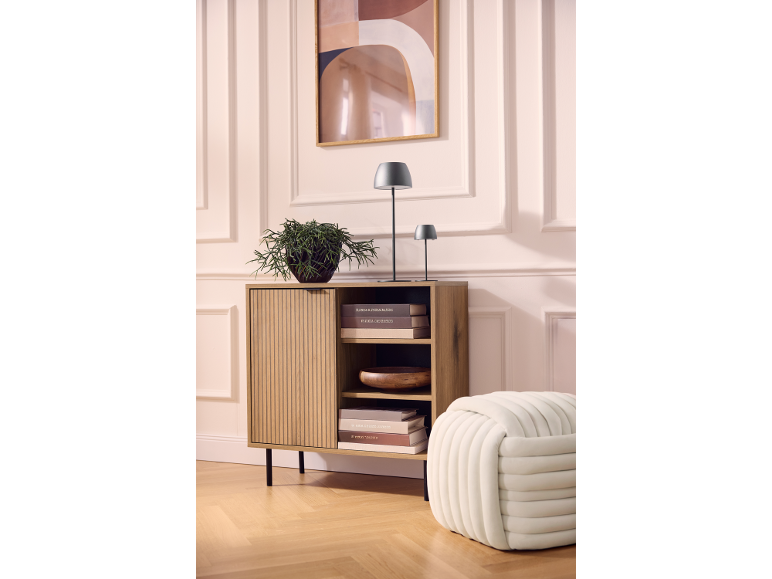 Wooden cabinet with a plant, lamps, and books, next to a pouf, against a wall with art.