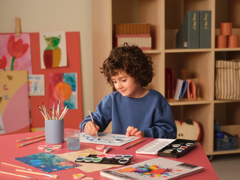 Child painting with watercolors and brushes on a pink table, with artwork in the background.