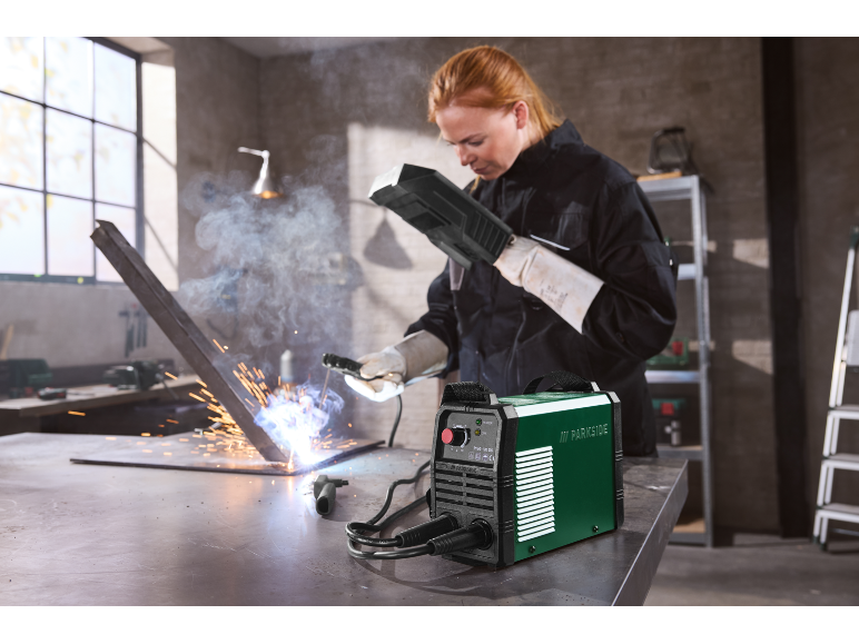 Woman in black overalls and protective gloves welding metal with a green welding machine.