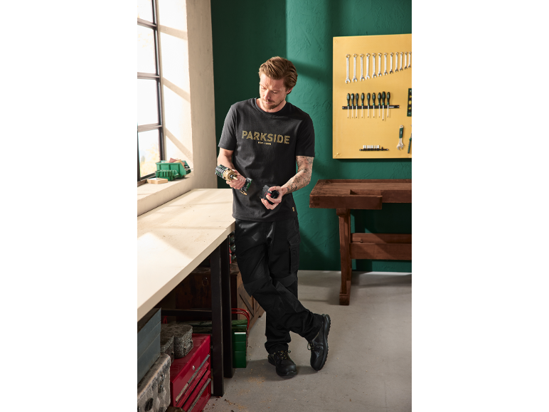 Man in black t-shirt and work trousers holding a cordless drill and battery in a workshop.