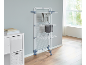 Vertical clothes drying rack with wheels, with clothes hanging, next to a white cabinet.