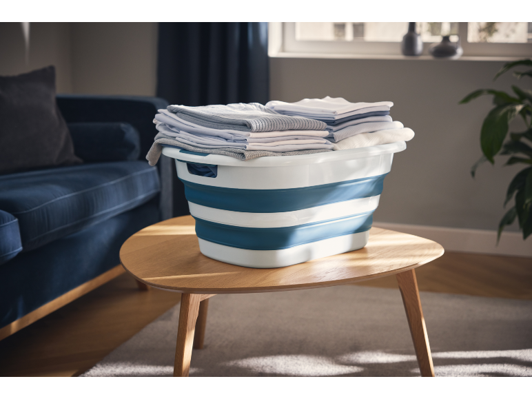 Folded laundry in a blue and white collapsible basket on a wooden table in a living room.