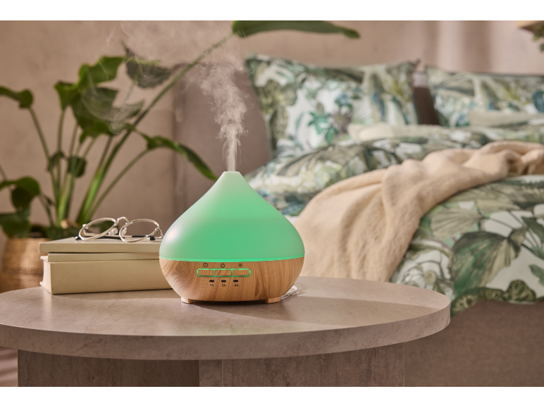 Green aroma diffuser with a wooden base in a bedroom with plants and patterned bedding.
