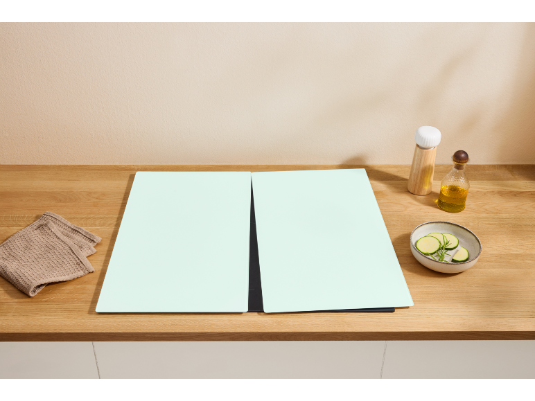 Wooden kitchen countertop with two light green cutting mats, a cloth, oil, and a bowl of zucchini.