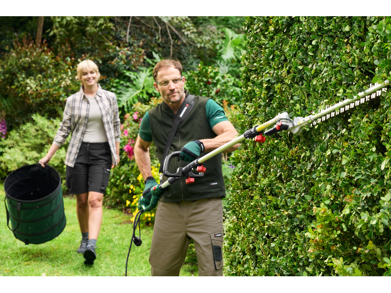 Man trimming hedge with a hedge trimmer, woman carrying a collapsible garden waste bag.