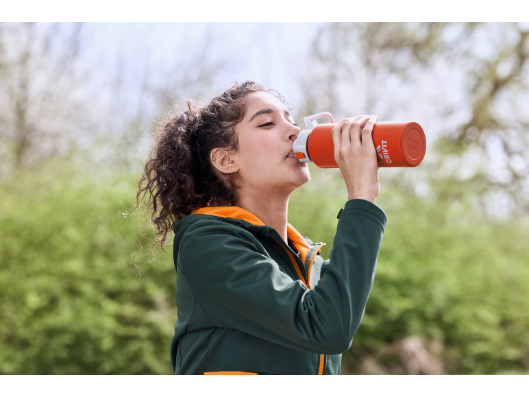 Young woman drinking from an orange water bottle, wearing a green jacket with an orange hoodie.