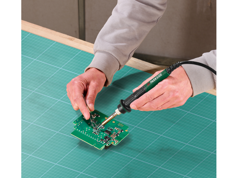 Man soldering a circuit board with a soldering iron and tweezers on a cutting mat.