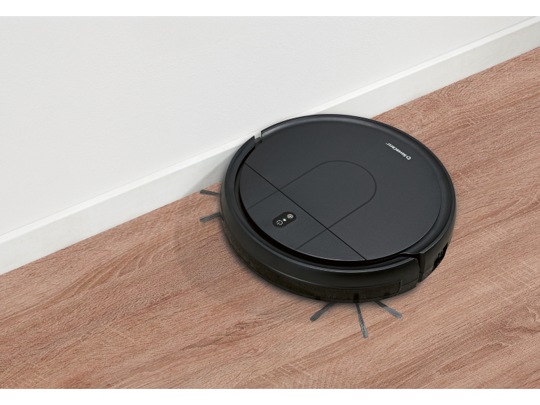 Black robotic vacuum cleaner cleaning a wooden floor next to a white wall.