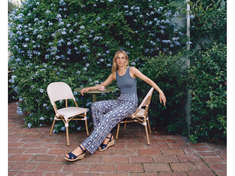 Woman in patterned trousers and wedge sandals sitting in a flowery patio.