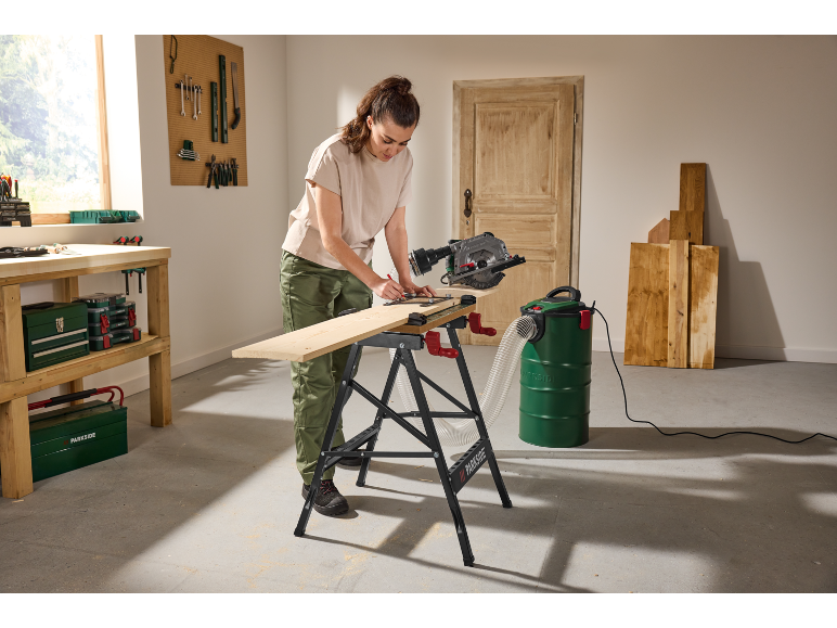 Woman using a circular saw and industrial vacuum cleaner to cut wood on a workbench.