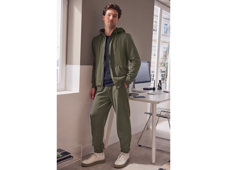 Man in olive green tracksuit set and sneakers, leaning against a wall.