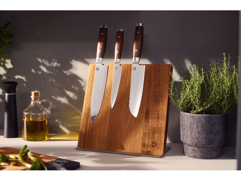 Wooden knife block with three knives, olive oil, pepper mill, and herb plant in a kitchen.