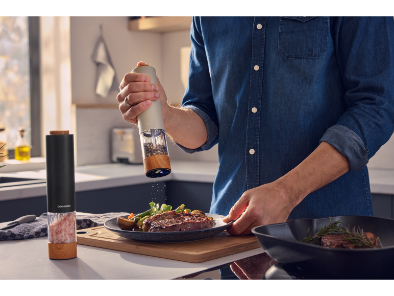 Man seasoning a steak with an electric pepper grinder in a kitchen.