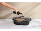 A hand pours red wine from a bottle into a roasting pan with a roasted chicken.
