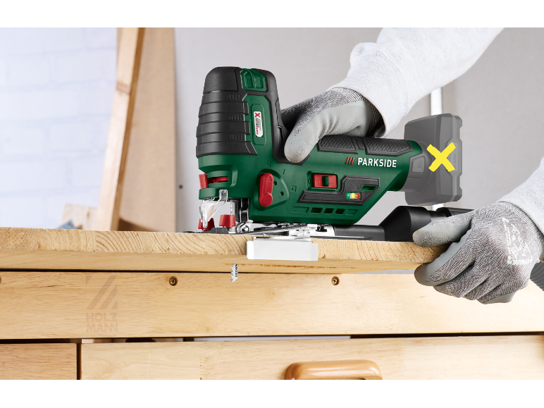 Gloved hands using a green jigsaw to cut a wooden board.