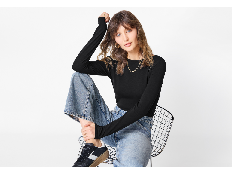Woman in black long-sleeve top, wide-leg jeans, and sneakers sitting on a chair.