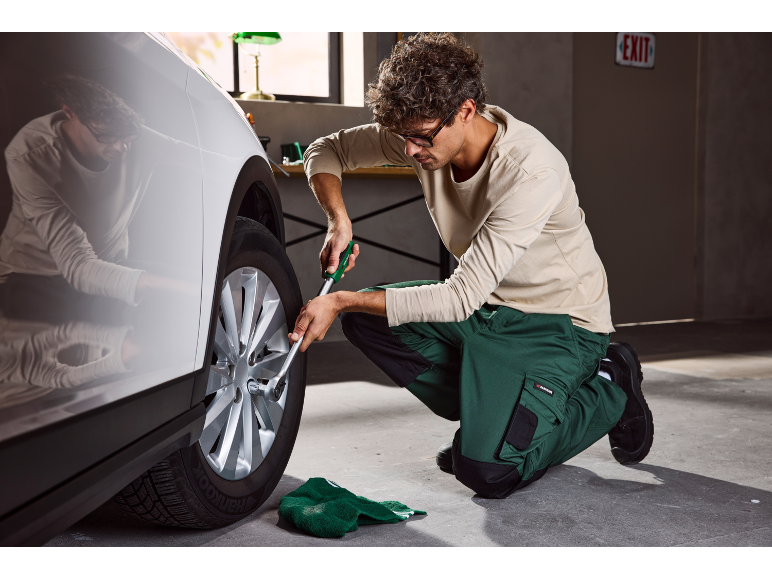 Man changing a tire with a lug wrench, wearing green work trousers.