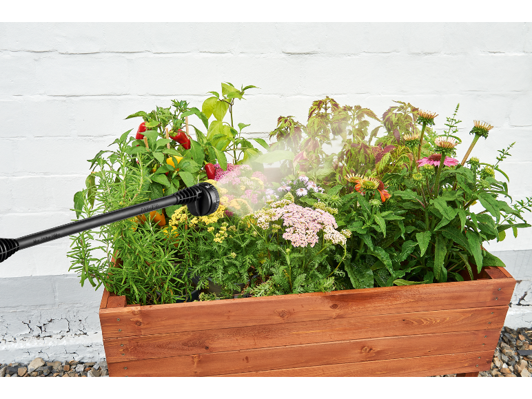 Hand spray nozzle watering a wooden raised garden bed with peppers, herbs, and flowers.