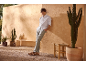 Man in white shirt and grey trousers, sandals, leaning against a sand-colored wall with cacti.