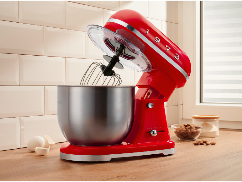 Red kitchen stand mixer with stainless steel bowl, eggs, and baking ingredients.