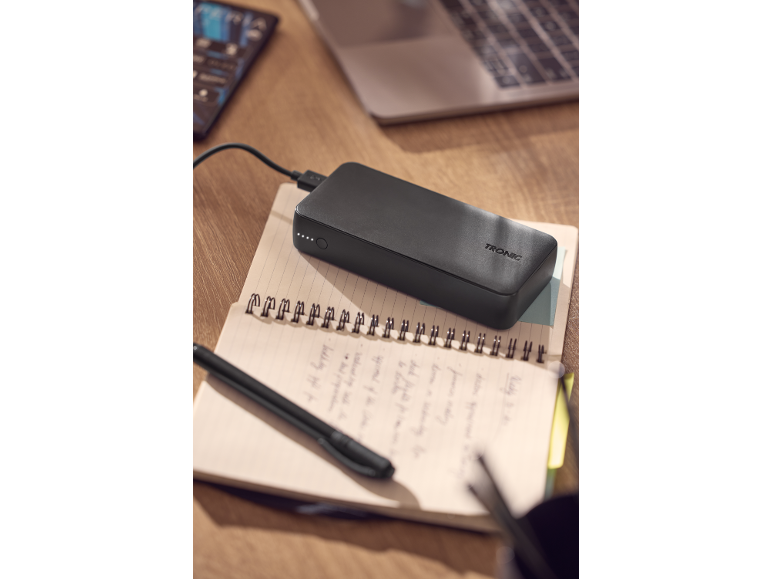 Black power bank charging on an open notebook with a pen, a laptop in the background.