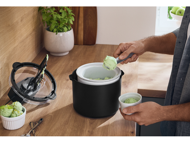 Man scooping ice cream from an ice cream maker into a cup on a wooden counter.