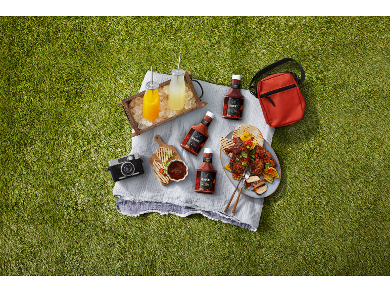 BBQ sauces, refreshing drinks, and grilled food at a picnic on grass.