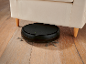 Black robotic vacuum cleaner cleaning under a beige sofa on a wooden floor.