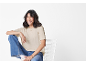 Smiling woman in a beige t-shirt and blue jeans, sitting on a white chair.