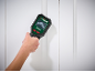 Hand holding a digital wall scanner against a white wall, displaying a signal.
