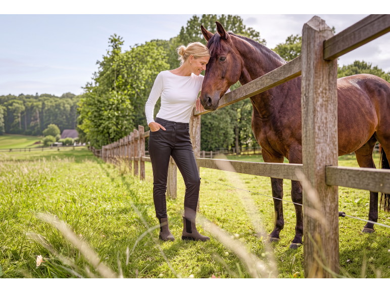Woman in black trousers and white top petting a brown horse by a wooden fence.