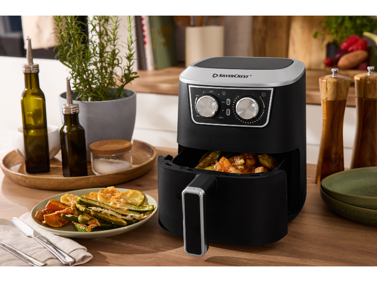 Air fryer with roasted vegetables, oil bottles, and spices in a kitchen setting.