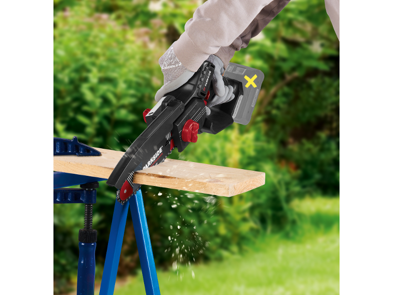 Gloved person using a cordless mini chainsaw to cut a wooden board outdoors.