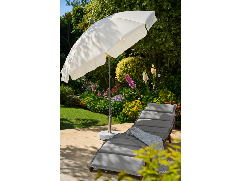 Sun lounger with grey cushion and white parasol on a patio by a flower garden.