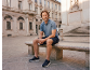 Man in blue polo shirt and dark shorts sitting on a stone bench outdoors.