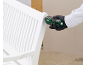A person in gloves uses a green electric screwdriver to assemble a white garden bench.
